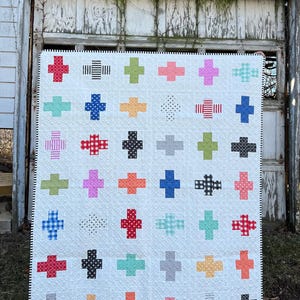 May include: A white quilt with a patchwork design featuring a variety of colorful plus signs. The plus signs are made from different fabrics with various patterns, including polka dots, stripes, gingham, and solid colors.