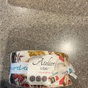 May include: A fabric bundle wrapped with a white ribbon and a tag that reads "Atelier 3 Sisters moda". The fabric has a floral pattern in shades of red, brown, and cream. The ribbon has the word "moda" printed on it.