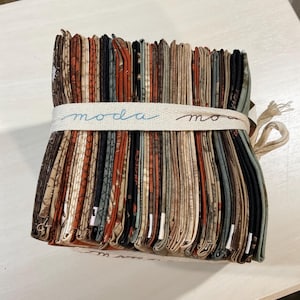 May include: A stack of fabric squares in various shades of brown, green, and black with a white fabric band that says "moda" on it.