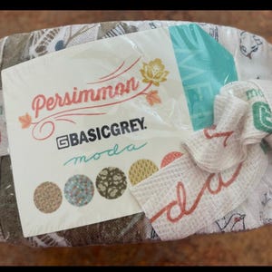 May include: A bundle of fabric squares wrapped in clear plastic. The label reads "Persimmon" and "BASICGREY moda." A small white towel with red script and a green design is tied around the bundle. The fabric has a floral and geometric pattern.