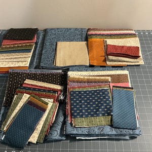 May include: A collection of assorted fabric squares and rectangular pieces in various colors including blue, brown, cream, orange, and red. The textiles feature diverse patterns such as small floral prints, dots, and solid textures, arranged in stacks.