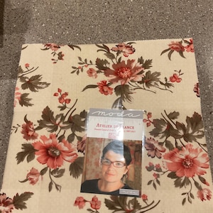 May include: A square fabric piece with a cream background and a large floral pattern in shades of red, pink, and brown. A small card with the text "Atelier de France" and a woman's portrait is on top of the fabric.