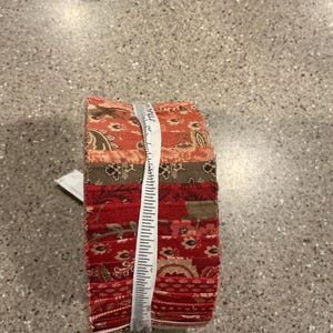 May include: A stack of patterned fabric squares in shades of red, brown, and tan, bound with a white measuring tape. The fabrics feature floral and paisley designs. The stack is approximately 3 inches tall.