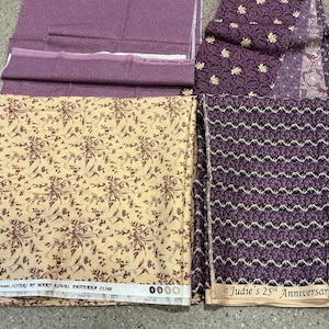 May include: A collection of folded cotton fabric pieces in shades of purple and cream. The fabrics feature various patterns including floral prints, small geometric shapes, and wavy lines. Selvedge text includes Windham Fabrics and Judie's 25th Anniversary.