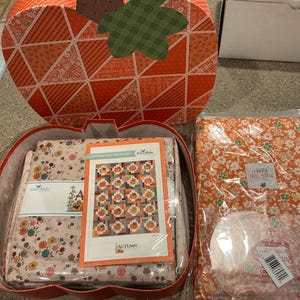 May include: An orange and brown pumpkin-shaped box with a quilt kit inside. The kit includes fabric with floral patterns, a pattern, and a clear plastic template. A second package of floral fabric and a white box are also visible.