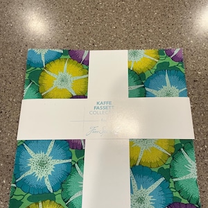 May include: A stack of fabric squares featuring a floral pattern with blue, yellow, and purple flowers on a green background. The fabric is wrapped with a white band that has text that reads "KAFFE FASSETT COLLECT" and the signature "Free Spirit".