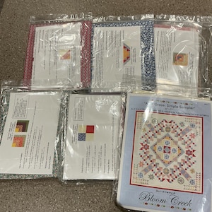 May include: A collection of quilting kits, each sealed in clear plastic. The kits feature various fabric patterns in red, blue, and green. One kit displays a finished quilt design with the text "At Grace: Simple Sampler" and "Bloom Creek".