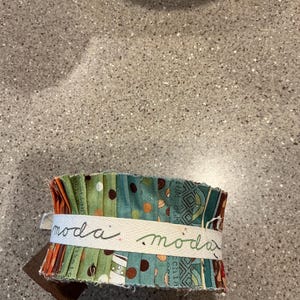 May include: A bundle of fabric strips in shades of green, teal, and orange, wrapped with a cream-colored ribbon. The ribbon is printed with the word "moda" in a handwritten style. A brown tag is attached.