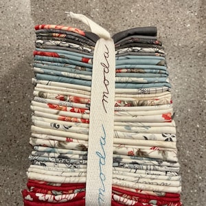 May include: A stack of folded fabric squares secured with a white ribbon printed with the word moda. The fabric features various patterns in shades of red, cream, light blue, and dark gray, including floral motifs and script text.