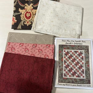 May include: A collection of five folded fabric squares in black, cream, tan, pink, and dark red, accompanied by a quilt pattern booklet titled Four Plus One Equals Fun: Jacob's Journey by Lori Smith, featuring a graphic of a finished quilt design.
