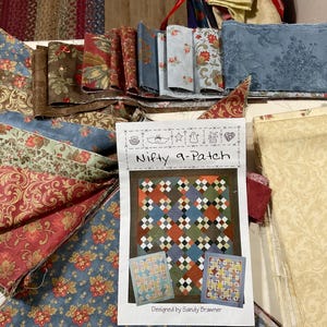 May include: A collection of fabric scraps in various patterns and colors, including red, blue, green, and brown. The fabrics are arranged in a semi-circle around a printed pattern for a quilt called "Nifty 9-Patch" designed by Sandy Brawner.