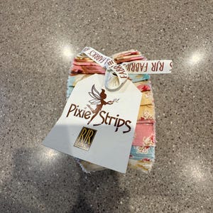 May include: A stack of fabric strips in pastel colors, tied with a ribbon that reads "RJR Fabrics." A white tag with the words "Pixie Strips" and a fairy illustration is attached to the fabric bundle.