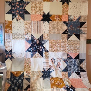 May include: A large, handmade quilt with a star pattern. The quilt features a variety of fabrics in shades of navy, peach, cream, and gold. The star shapes are prominent, and the quilt has a patchwork design. The quilt is hanging on a wall.