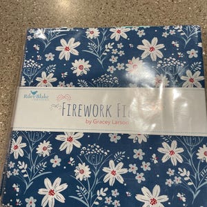 May include: A square stack of fabric squares wrapped in clear plastic, featuring a dark blue background with a white and red floral pattern. A white paper band across the center displays the text Riley Blake Designs, Firework Fiesta by Gracey Larson.