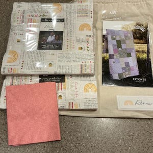 May include: A collection of fabric bundles and a pattern for a quilt. The fabric bundles are wrapped in clear plastic and feature a cream-colored background with text and rainbow designs. A small pink checkered fabric square is also included.