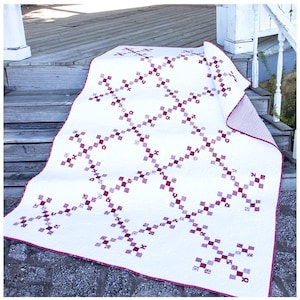 May include: A white quilt with a repeating pattern of purple and burgundy squares and floral motifs. The quilt has a burgundy border and is displayed on wooden steps. The reverse side of the quilt has a white and burgundy polka dot pattern.