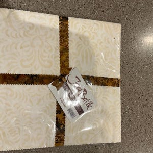 May include: A stack of cream-colored fabric squares with a subtle floral pattern, bound with a brown patterned ribbon. A tag reading "Java Batik Studio" is attached. The fabric appears to be packaged in clear plastic.