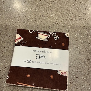 May include: A stack of brown fabric squares with coffee-themed patterns, including coffee cups and beans. The fabric is wrapped with a white band that reads "moda JAVA by DEB STRAIN for moda."