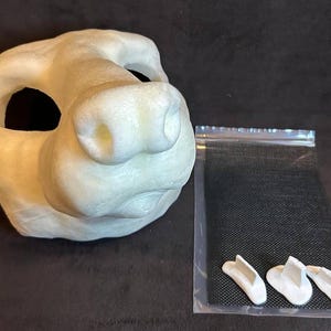 May include: A light beige, unfinished animal mask with large eye openings and a prominent snout. Next to the mask is a small plastic bag containing three white, curved pieces, possibly for the mask's construction. The items are on a dark surface.