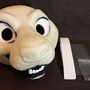 May include: A white foam fursuit head with black eyes and a black nose. The head is open-mouthed with white teeth. The head is accompanied by a white strap, black mesh fabric, and a plastic bag containing white foam pieces.