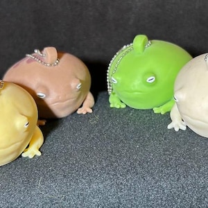 Puede incluir: Cuatro pequeñas figuritas con forma de rana con cuerpos de diferentes colores. Las ranas están hechas de un material suave y blando y tienen una cadena unida a la parte superior de sus cabezas. Las ranas están sentadas sobre una superficie oscura.