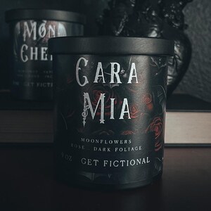 May include: Two black candle jars with white and red labels. The candle on the left says "Mon Cheri" and the candle on the right says "Gara Mia" with the text "Rose, Moonflowers, Dark Foliage" and "10oz Get Fictional".