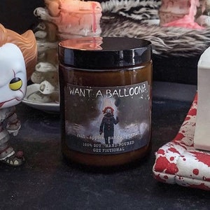 May include: A brown glass candle with a black lid and a label that says "Want a Balloon?" with a picture of Pennywise the clown from the movie "IT". The candle is sitting on a table next to a figurine of Pennywise the clown and a book by Stephen King.