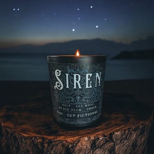 May include: A lit candle in a black glass jar with the word "SIREN" in white script. The jar has a decorative pattern and the words "SALT, SEA MOSS, BLACK PLUM, AMBER" and "GET FICTIONAL". The candle sits on a wooden surface with a night sky background.