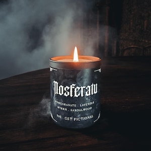 May include: A black candle with a white label that reads "Nosferatu" and "Pomegranate, Lavender, Myrrh, Sandalwood, 9oz. Get Fictional". The candle is lit and smoke is rising from the top.