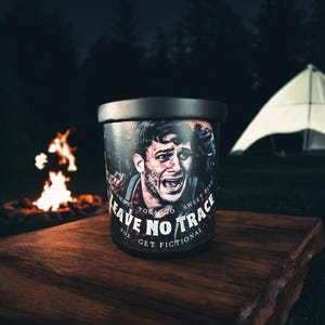 May include: A black and silver can with a graphic of a man's face and the text "Leave No Trace" in white. The can is sitting on a wooden table in front of a campfire and a tent.