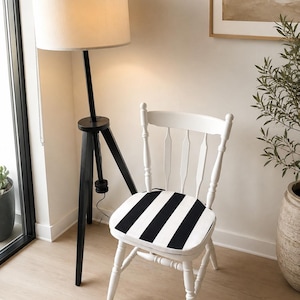 May include: A white wooden chair with a seat cushion featuring bold black and white vertical stripes, positioned next to a black tripod floor lamp with a cylindrical off-white fabric lampshade that emits a soft, warm light.