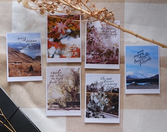 May include: A collection of six rectangular postcards with nature-themed photographs and inspirational quotes in cursive script. The images feature landscapes, flowers, and trees. The postcards are arranged on a checkered surface.