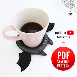 Może przedstawiać: Jasnoróżowy kubek ceramiczny stoi na ciemnoszarej, materiałowej podstawce w kształcie nietoperza z wyhaftowanymi detalami. Tekst: YouTube tutorials + PDF SEWING PATTERN. Mały czarny pająk wisi na pajęczynie w prawym górnym rogu.
