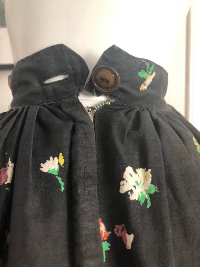 May include: A black skirt featuring a floral print with white, yellow, and red flowers. The waistband includes a single brown button closure, a horizontal buttonhole, and a metal zipper located directly below the waistband center.