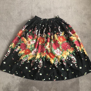 May include: A black knee-length skirt featuring a vibrant floral print with clusters of red, orange, yellow, and white flowers. The skirt has a gathered waistband with pleats that create a full, flared silhouette and a simple hemline.