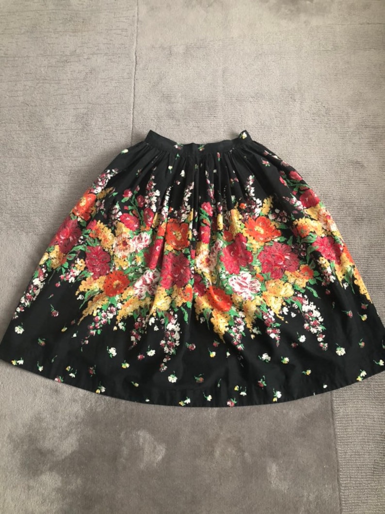 May include: A black knee-length skirt featuring a vibrant floral print with clusters of red, orange, yellow, and white flowers. The skirt has a gathered waistband with pleats that create a full, flared silhouette and a simple hemline.