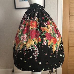 May include: A black, high-waisted, full-length skirt featuring a vibrant, multi-colored floral print with red, yellow, and green blossoms. The skirt has a voluminous, pleated silhouette that flares out from the waist, creating a structured shape.