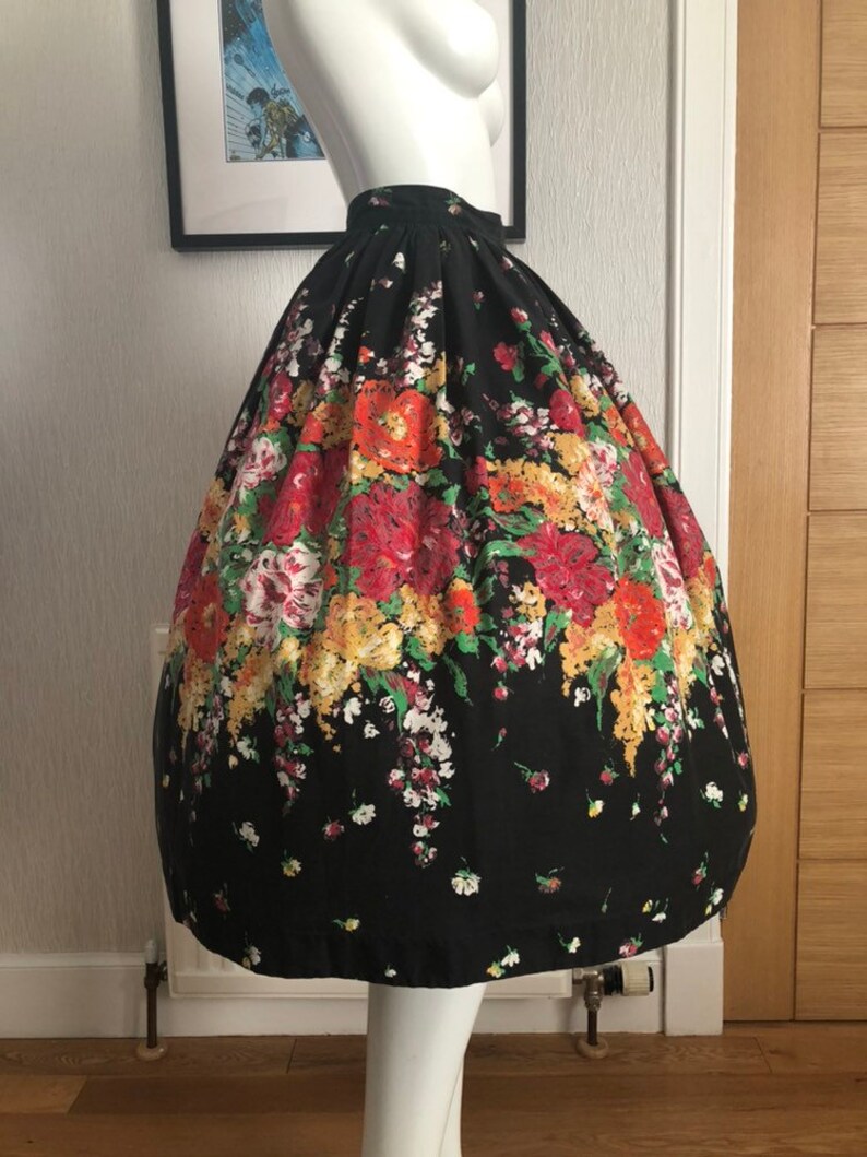 May include: A black, high-waisted, full-length skirt featuring a vibrant, multi-colored floral print with red, yellow, and green blossoms. The skirt has a voluminous, pleated silhouette that flares out from the waist, creating a structured shape.