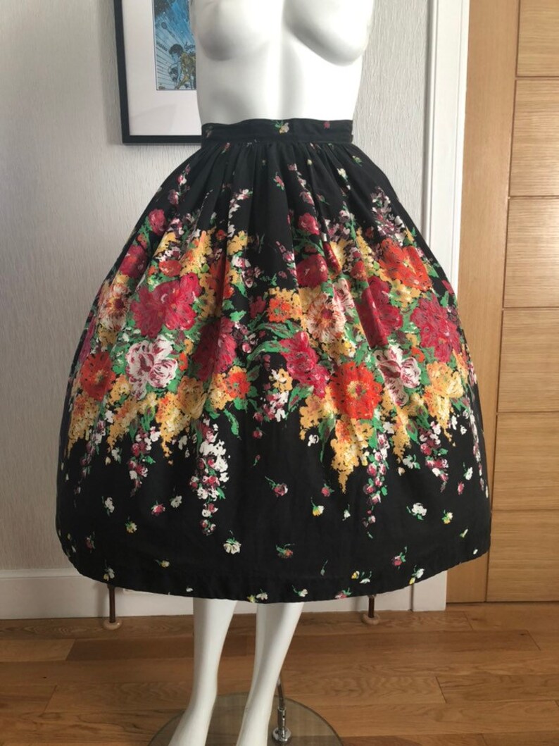 May include: A high-waisted, full-length skirt featuring a black base color with a vibrant floral print. The pattern includes clusters of red, orange, yellow, and white flowers with green foliage. The skirt has a gathered waistband and a voluminous silhouette.