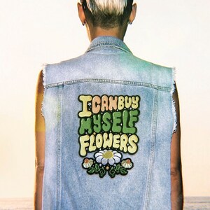 Peut inclure: Une personne portant un gilet en jean bleu clair avec un patch qui dit "Je peux m'acheter des fleurs" en lettres colorées avec un motif de marguerite.