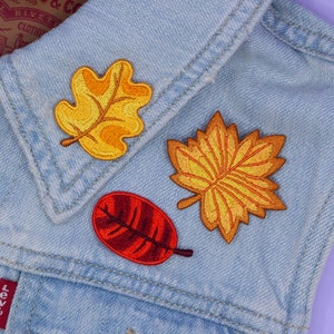Pode incluir: Três patches bordados para passar a ferro com motivos de folhas de outono em tons de vermelho, laranja e amarelo. Os patches estão sobre uma jaqueta de ganga azul.