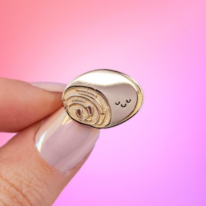 Peut inclure: Épinglette dorée en forme d'escargot de dessin animé avec une coquille en spirale et un petit visage aux yeux fermés.