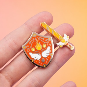 May include: A set of two enamel pins featuring a shield with a flame design and a sword with wings. The shield is orange with gold accents and the sword is yellow with gold accents.