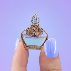Peut inclure: Une épingle en émail dorée en forme de flacon avec un liquide bleu et une couronne dorée. L'épingle est tenue par un doigt.