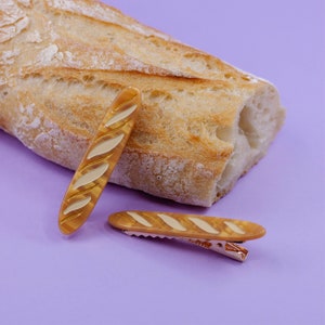May include: Two brown and beige baguette-shaped hair clips on a purple background. One clip is laying horizontally and the other is standing vertically.