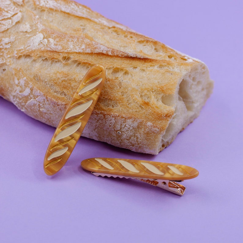May include: Two brown and beige baguette-shaped hair clips on a purple background. One clip is laying horizontally and the other is standing vertically.