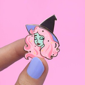K&ouml;nnte beinhalten: Eine rosa und gr&uuml;ne Hexen-Emaille-Anstecknadel mit einem schwarzen Hexenhut und einem glitzernden Design.