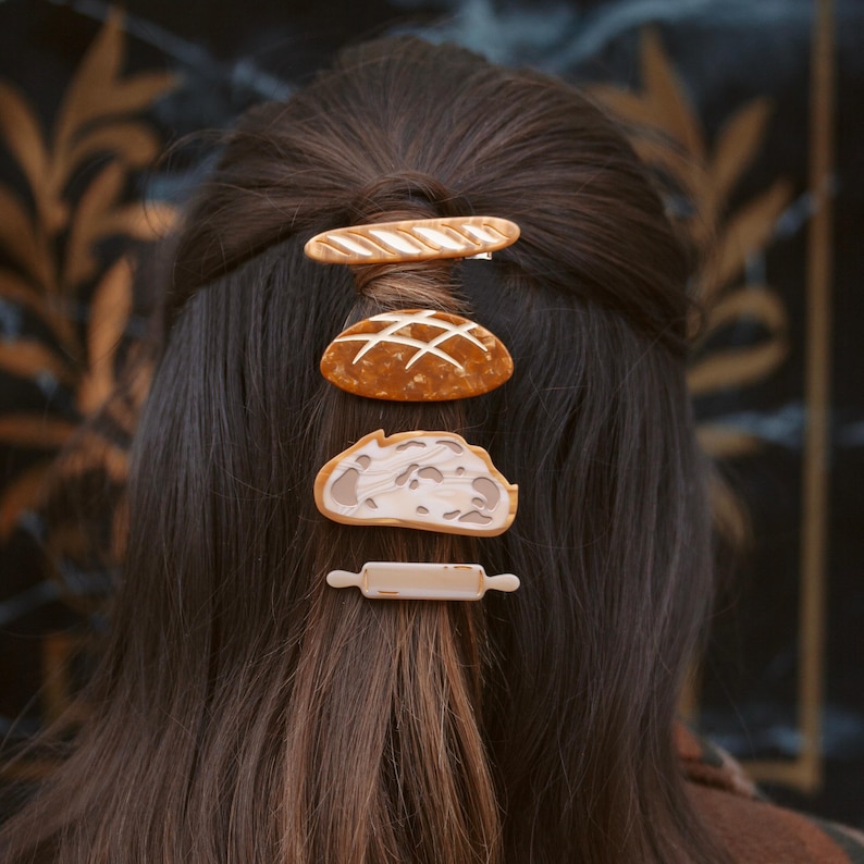 May include: Four hair clips in the shape of bread and a rolling pin. The clips are brown and beige and are arranged in a vertical line on a person's brown hair.