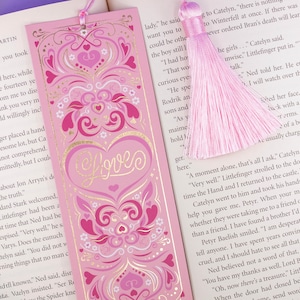 Può includere: Un segnalibro rosa con un design a forma di cuore dorato e la parola "Love" in lettere dorate. Il segnalibro ha un fiocco rosa.
