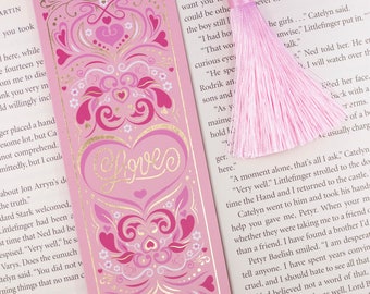 Peut inclure: Un marque-page rose avec un motif de cœur doré et le mot "Love" en lettres dorées. Le marque-page a un pompon rose.